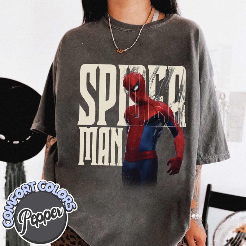 Peut inclure: T-shirt gris fonc&eacute; avec un graphisme de Spider-Man en costume rouge et bleu, et l'inscription "SPIDER MAN" en blanc. Le t-shirt a un aspect vieilli et le nom de la marque "Comfort Colors Pepper" est visible.
