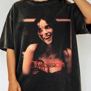 May include: A charcoal grey acid-wash t-shirt featuring a graphic print of a smiling person with dark hair and blood-like markings on their face. The word Obsession is printed in bold red letters at the bottom of the graphic design on the front.