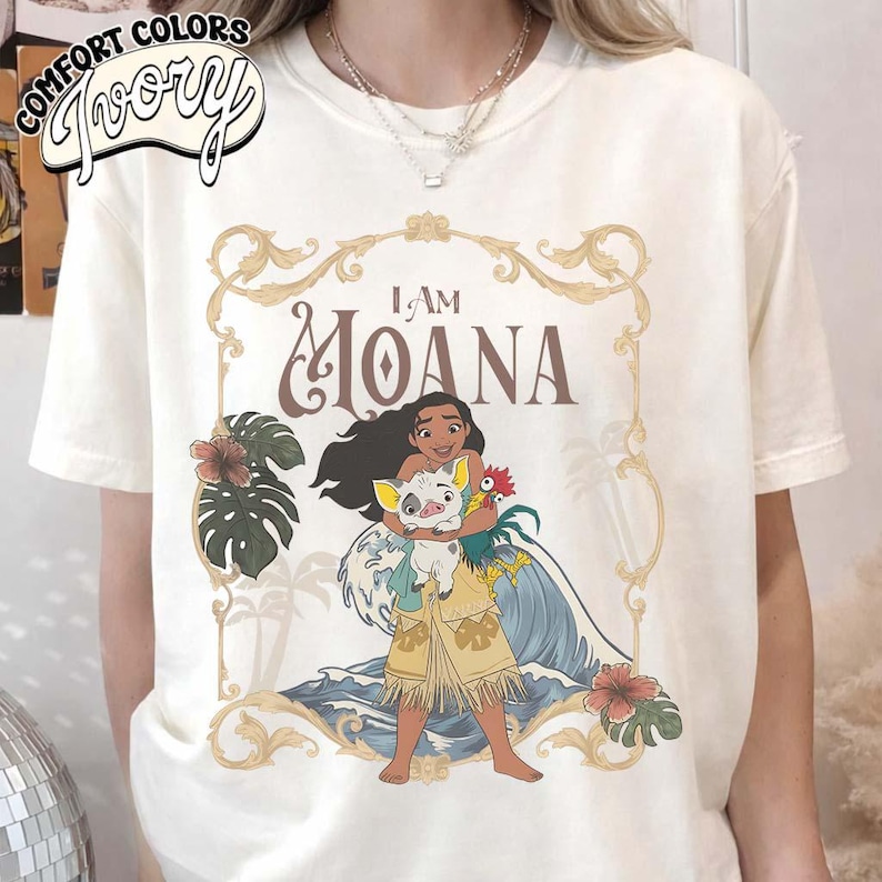 May include: An ivory t-shirt featuring a graphic print of the character Moana holding a pig and a rooster. The design includes the text I AM MOANA, surrounded by a gold ornate frame, tropical leaves, hibiscus flowers, and ocean waves.