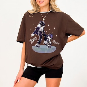Pu&ograve; includere: Una t-shirt a maniche corte color marrone scuro con una stampa grafica di Michael Jackson e Mickey Mouse su una piattaforma luminosa. Il design include accenti a forma di stella bianca e il testo Michael Jackson e Mickey Mouse in corsivo.