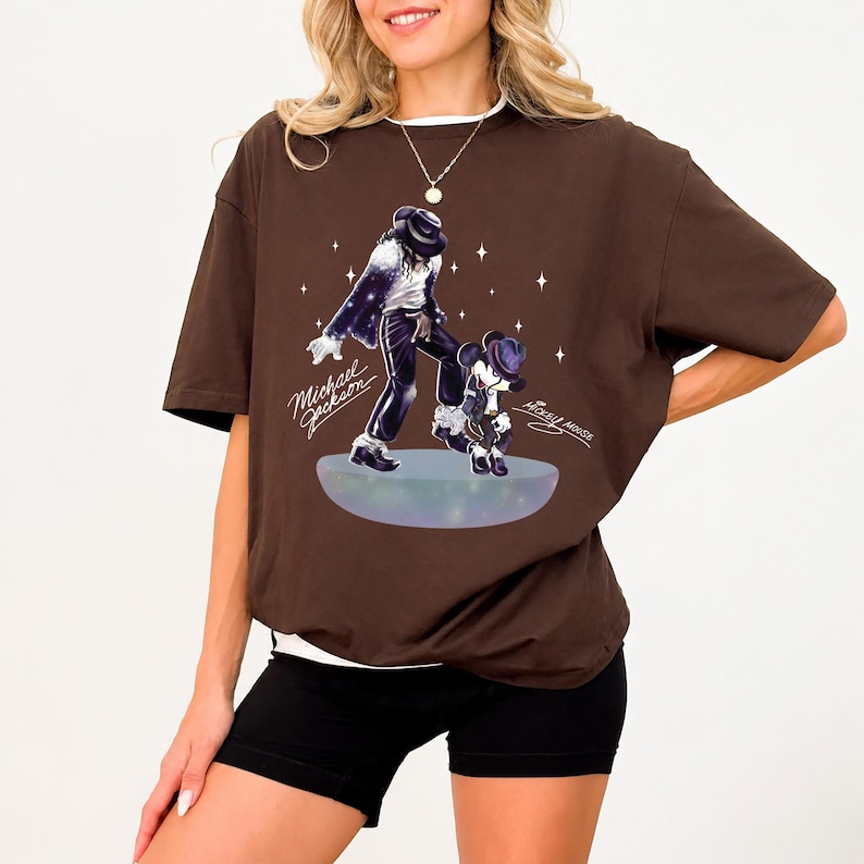 Pu&ograve; includere: Una t-shirt a maniche corte color marrone scuro con una stampa grafica di Michael Jackson e Mickey Mouse su una piattaforma luminosa. Il design include accenti a forma di stella bianca e il testo Michael Jackson e Mickey Mouse in corsivo.
