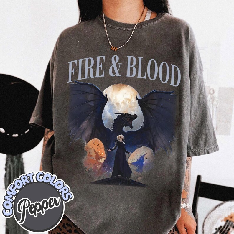 May include: A charcoal gray short-sleeve t-shirt featuring the text FIRE & BLOOD in a light blue font. The graphic depicts a large black dragon with spread wings standing behind a figure in a long black dress against a full moon background.