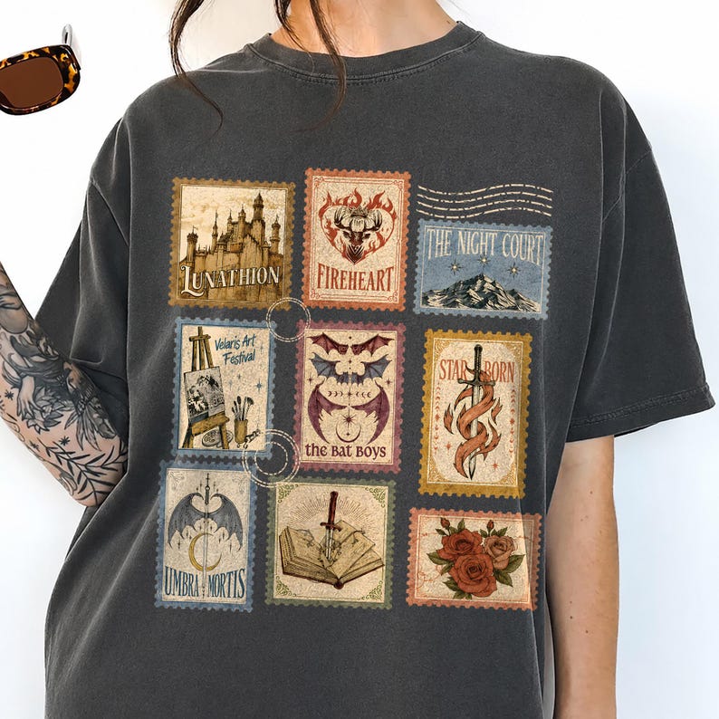 May include: A charcoal gray t-shirt featuring a grid of nine postage stamp-style illustrations. The designs include a castle labeled Lunathion, a deer head, a mountain range, an easel, bat wings, a sword, a book, and roses with text.