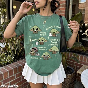 K&ouml;nnte beinhalten: Hellgr&uuml;nes T-Shirt mit Cartoon-Charakter-Illustrationen und S&auml;tzen wie "Keeping Strong" und "Having a Secret Weapon". Das Shirt hat auch den Text "Cardio" und "Sleeping 8 hours."