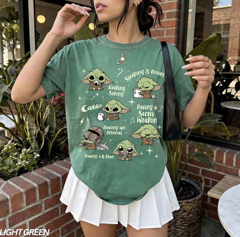 K&ouml;nnte beinhalten: Hellgr&uuml;nes T-Shirt mit Cartoon-Charakter-Illustrationen und S&auml;tzen wie "Keeping Strong" und "Having a Secret Weapon". Das Shirt hat auch den Text "Cardio" und "Sleeping 8 hours."