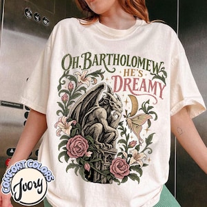May include: An ivory t-shirt featuring a graphic print of a gargoyle perched on a pedestal, surrounded by pink roses, white lilies, and a moth. The text reads "OH, BARTHOLOMEW HE'S DREAMY" in dark green and pink lettering with a crescent moon.
