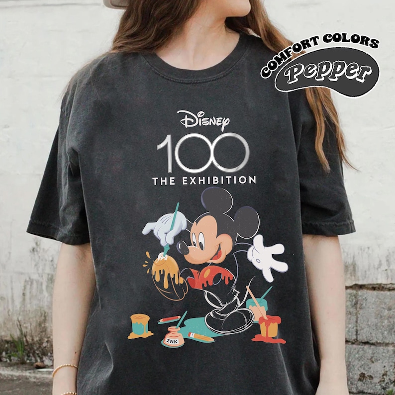 May include: Dark gray t-shirt featuring Mickey Mouse painting with paint pots and the text "Disney 100 The Exhibition." The shirt also has the words "Comfort Colors Pepper" in a circular design.