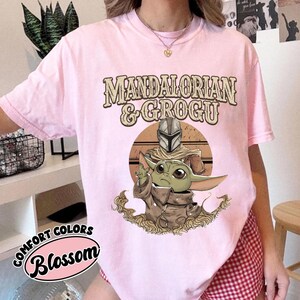 Puede incluir: Camiseta rosa claro con el texto "MANDALORIAN & GROGU" sobre una ilustraci&oacute;n de Mandalorian y Grogu. La camiseta tambi&eacute;n tiene las palabras "COMFORT COLORS" y "Blossom" en un dise&ntilde;o circular.