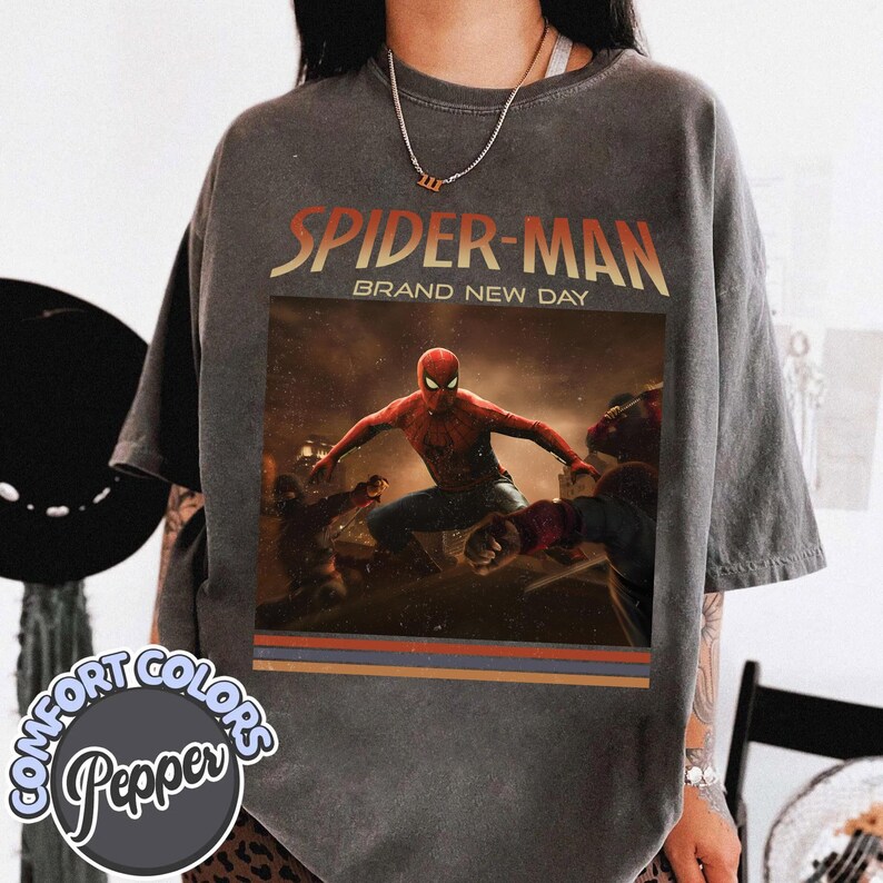 May include: A charcoal gray short-sleeve t-shirt featuring a graphic print of Spider-Man in a dynamic action pose. The text "SPIDER-MAN" and "BRAND NEW DAY" appear above the character. The design includes horizontal stripes at the bottom.