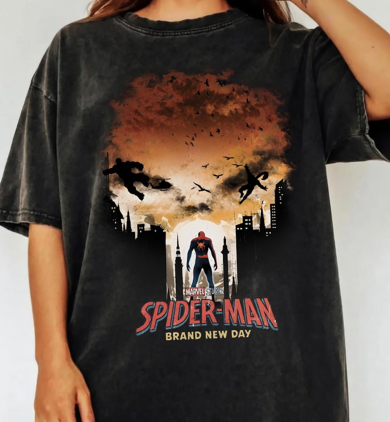 Może przedstawiać: Ciemnoszara koszulka z grafiką Spider-Mana. Projekt zawiera sylwetki postaci, krajobrazy miejskie oraz napis "SPIDER-MAN BRAND NEW DAY" w kolorze czerwonym i niebieskim. Widoczne jest r&oacute;wnież logo Marvel Studios.