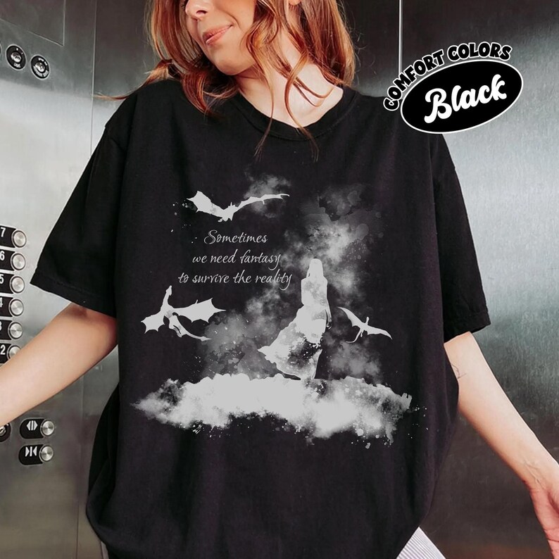 May include: A black short-sleeve t-shirt featuring a white graphic of a silhouette standing amidst clouds with three flying dragons. The text reads, Sometimes we need fantasy to survive the reality. A circular badge in the corner says Comfort Colors Black.