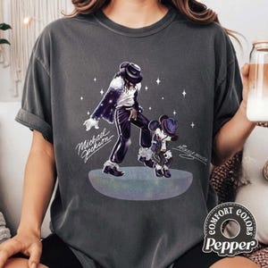 Pu&ograve; includere: Una t-shirt grigio antracite a maniche corte con una grafica di Michael Jackson e Mickey Mouse che ballano su una piattaforma circolare. Il design include stelle bianche e il testo Michael Jackson e Mickey Mouse in corsivo.