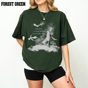 May include: A forest green short-sleeved t-shirt featuring a white graphic print of a silhouette, two flying dragons, and the text: Sometimes we need fantasy to survive the reality. The shirt has a crew neckline and a loose, relaxed fit.