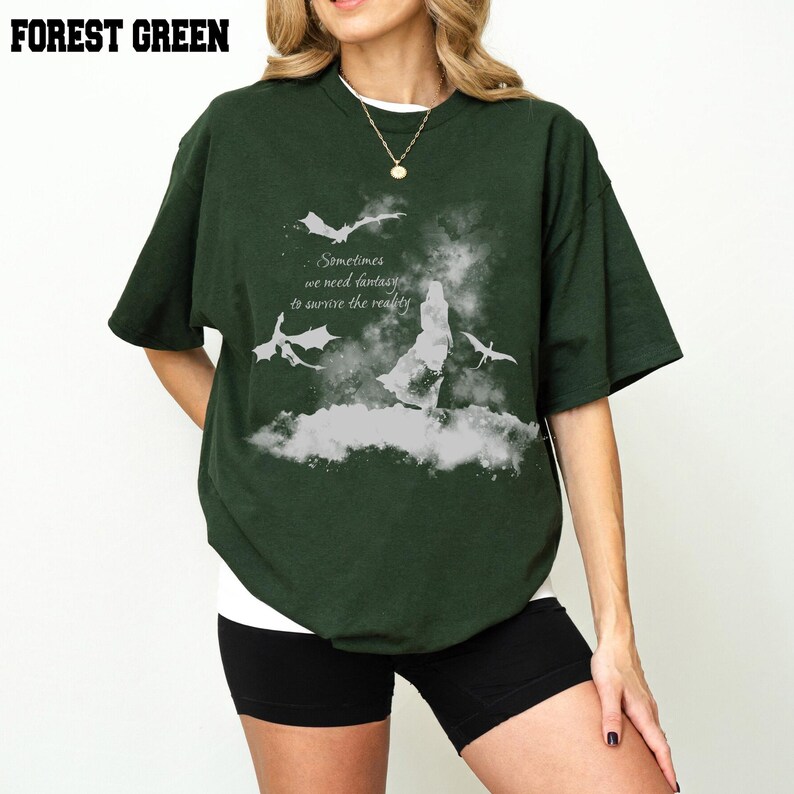 May include: A forest green short-sleeved t-shirt featuring a white graphic print of a silhouette, two flying dragons, and the text: Sometimes we need fantasy to survive the reality. The shirt has a crew neckline and a loose, relaxed fit.