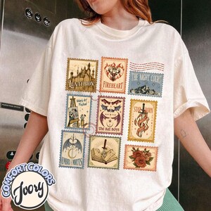 May include: An ivory t-shirt featuring a grid of nine rectangular postage stamp-style graphics. The designs include a castle, a deer, a mountain, an easel, bat wings, a sword, a dagger, an open book, and red roses with various text labels.