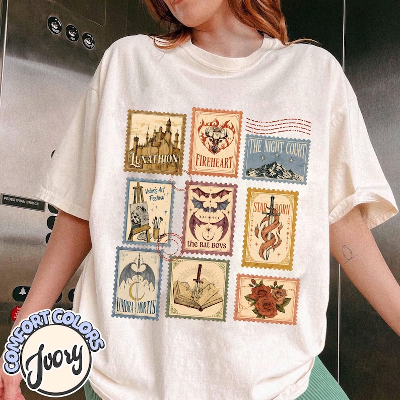 May include: An ivory t-shirt featuring a grid of nine rectangular postage stamp-style graphics. The designs include a castle, a deer, a mountain, an easel, bat wings, a sword, a dagger, an open book, and red roses with various text labels.