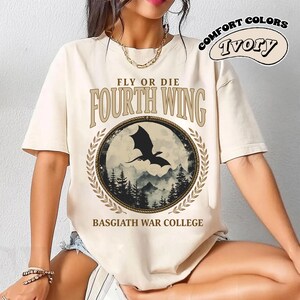 Puede incluir: Camiseta de color marfil con el texto "FLY OR DIE FOURTH WING" sobre un gr&aacute;fico circular de una silueta de drag&oacute;n sobre un paisaje monta&ntilde;oso. Las palabras "BASGIATH WAR COLLEGE" est&aacute;n debajo. La camiseta tiene mangas cortas y un corte holgado.