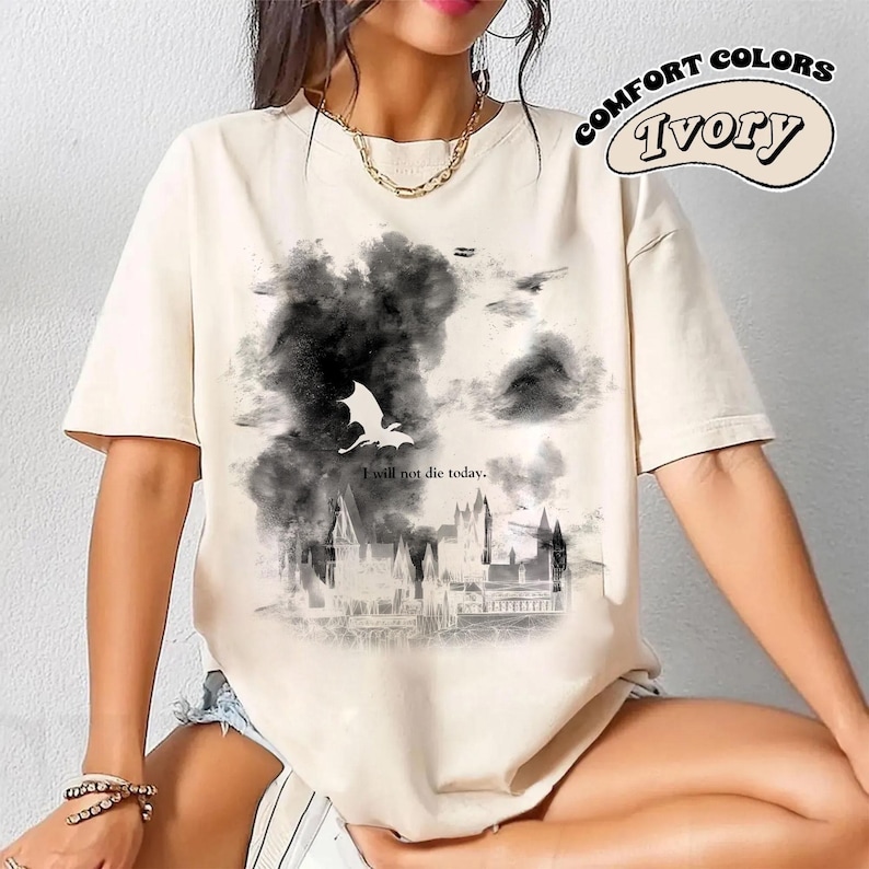 May include: Cream-coloured t-shirt with a graphic design featuring a dragon, dark clouds, and a castle. The text "I will not die today" is below the dragon. The words "Comfort Colors" and "Ivory" are in a circular design.