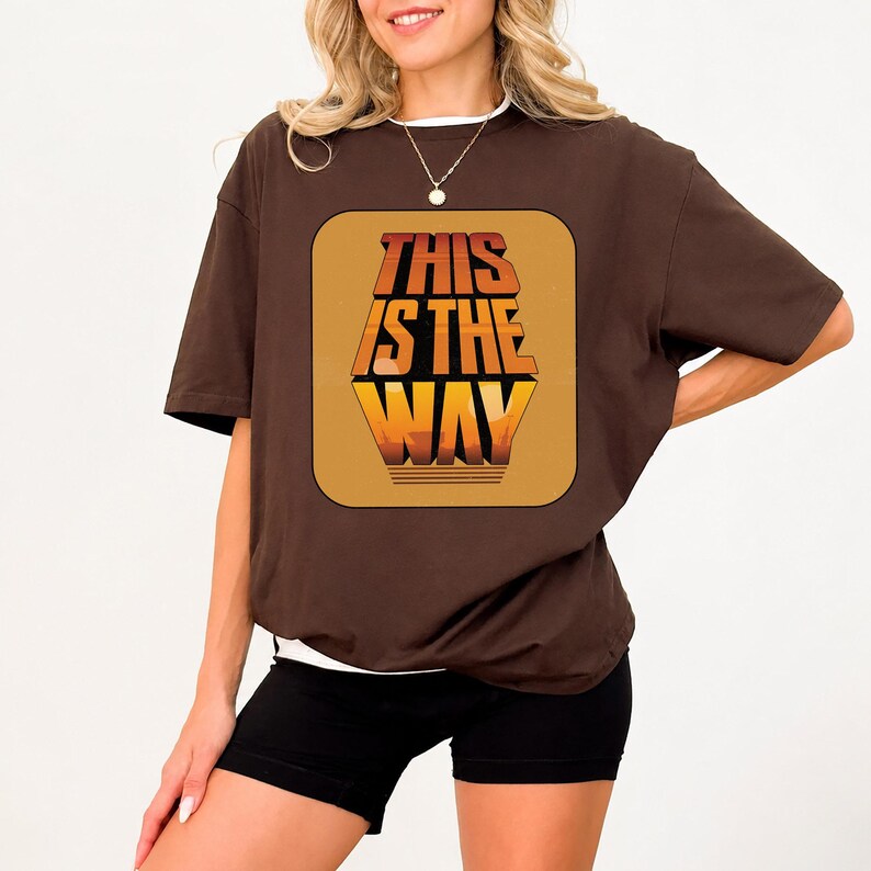 K&ouml;nnte beinhalten: Ein braunes T-Shirt mit dem Text "THIS IS THE WAY" in einem Retro-Schriftstil. Der Text ist in Orange- und Gelbt&ouml;nen gehalten und befindet sich auf einem braunen Quadrat. Das Shirt hat kurze &Auml;rmel und einen wei&szlig;en Kragen.