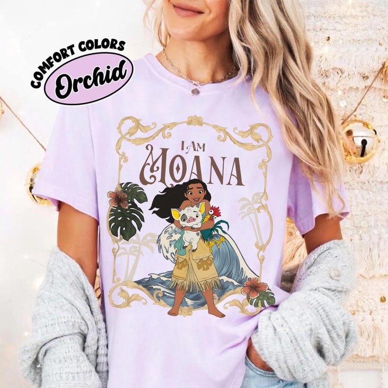 May include: A light purple short-sleeve t-shirt featuring a graphic of the character Moana holding a pig and a rooster. The design includes the text I AM MOANA inside a gold ornate frame with tropical leaves and ocean waves in the background.