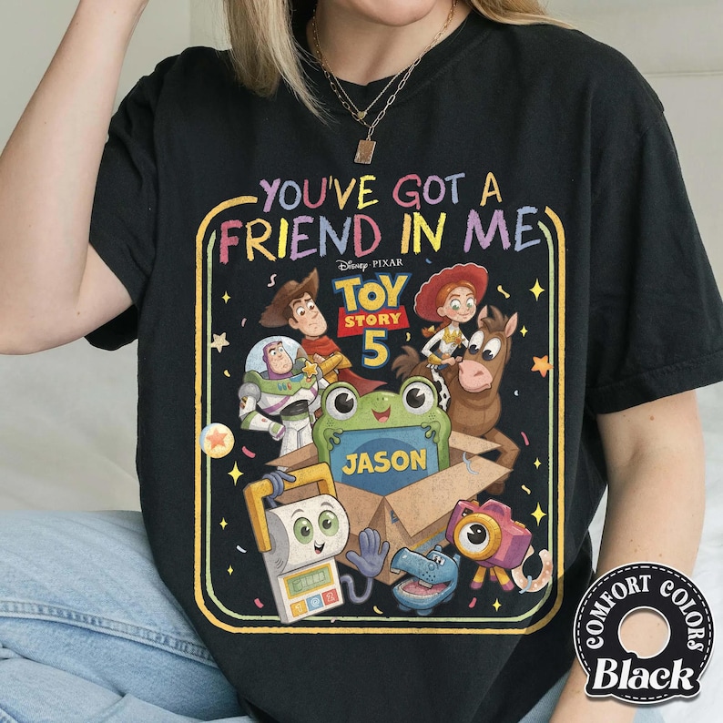 May include: A black t-shirt featuring a colorful graphic with the text "YOU'VE GOT A FRIEND IN ME" and "TOY STORY 5". The design includes illustrations of Woody, Buzz Lightyear, Jessie, Bullseye, and a green frog holding a box labeled "JASON".