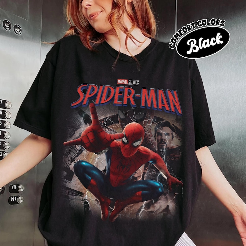 May include: A black short-sleeve t-shirt featuring a graphic print of Spider-Man in a web-shooting pose. Above the character is the red and blue text Spider-Man and the Marvel Studios logo. A small oval badge in the corner reads Comfort Colors Black.
