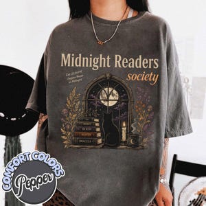 May include: A charcoal gray t-shirt featuring a graphic of a black cat sitting before an arched window with a full moon. The text reads Midnight Readers society, Est. 11:59 PM, Chapter Begun at Midnight, and includes book titles Poe, Bronte, Dickens, Austen, Dracula.