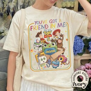 May include: An ivory t-shirt featuring a colorful graphic with the text "YOU'VE GOT A FRIEND IN ME" and "TOY STORY 5". The design includes illustrations of Woody, Buzz Lightyear, Jessie, Bullseye, and other characters around a box labeled "JASON".