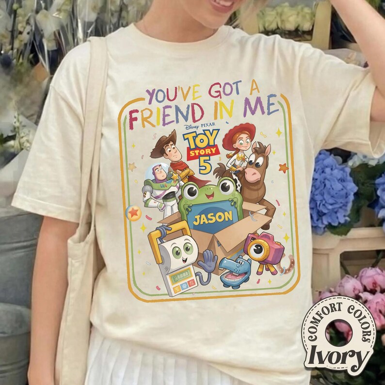 May include: An ivory t-shirt featuring a colorful graphic with the text "YOU'VE GOT A FRIEND IN ME" and "TOY STORY 5". The design includes illustrations of Woody, Buzz Lightyear, Jessie, Bullseye, and other characters around a box labeled "JASON".