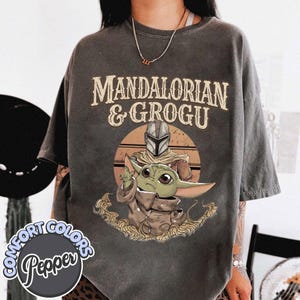 Puede incluir: Camiseta gris oscuro con el texto "MANDALORIAN & GROGU" sobre una ilustraci&oacute;n del Mandaloriano y Grogu. La ilustraci&oacute;n muestra a Grogu con una capa marr&oacute;n y al Mandaloriano con su casco. La camiseta tiene el logo Comfort Colors Pepper.