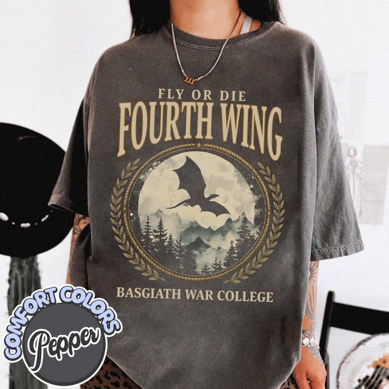 Puede incluir: Camiseta gris oscuro con el texto "FLY OR DIE FOURTH WING" sobre un gr&aacute;fico circular de una silueta de drag&oacute;n sobre un paisaje monta&ntilde;oso. El texto "BASGIATH WAR COLLEGE" est&aacute; debajo. La camiseta es de la marca Comfort Colors.