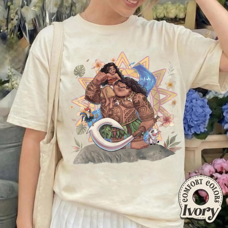 May include: An ivory short-sleeve t-shirt featuring a graphic print of Moana and Maui from the Disney film. The design includes Moana sitting on Maui's shoulders, a large white hook, a rooster, a pig, and a yellow sunburst background with flowers.