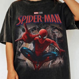 May include: A charcoal grey short-sleeve t-shirt featuring a graphic print of Spider-Man in a dynamic pose with web-shooting hands. The design includes the text Marvel Studios and Spider-Man in red and blue, alongside an image of Doctor Strange.
