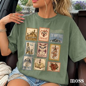 May include: An olive green short-sleeve t-shirt featuring a grid of nine rectangular postage stamp-style graphics. The designs include a castle, a deer head, a mountain range, an easel, bat wings, a sword, a crescent moon, a book, and roses.