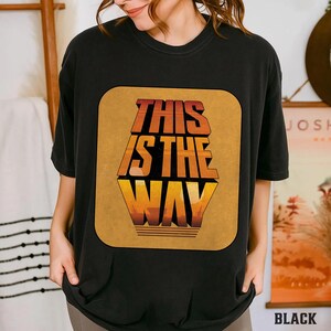 K&ouml;nnte beinhalten: Schwarzes T-Shirt mit einem gro&szlig;en, rechteckigen Grafikdesign. Das Design zeigt den Text "THIS IS THE WAY" in Orange und Gelb, mit einer Landschaft am unteren Rand. Das Shirt hat einen l&auml;ssigen Stil.