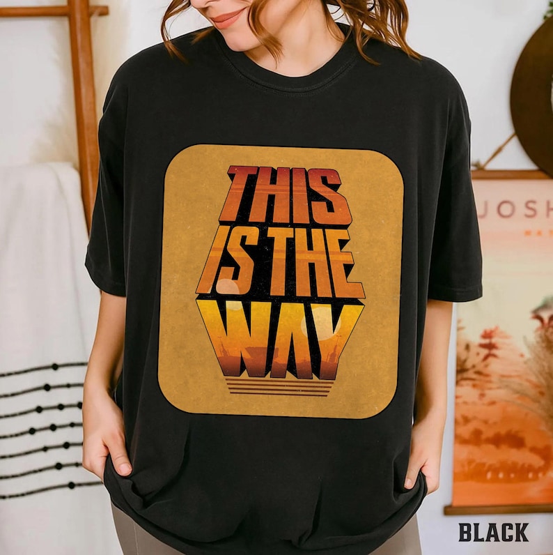 K&ouml;nnte beinhalten: Schwarzes T-Shirt mit einem gro&szlig;en, rechteckigen Grafikdesign. Das Design zeigt den Text "THIS IS THE WAY" in Orange und Gelb, mit einer Landschaft am unteren Rand. Das Shirt hat einen l&auml;ssigen Stil.
