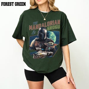 May include: Forest green t-shirt featuring a graphic of The Mandalorian and Grogu, with a spaceship and a planet in the background. The text "THE MANDALORIAN & GROGU" is printed above the graphic. The shirt is a dark green color.