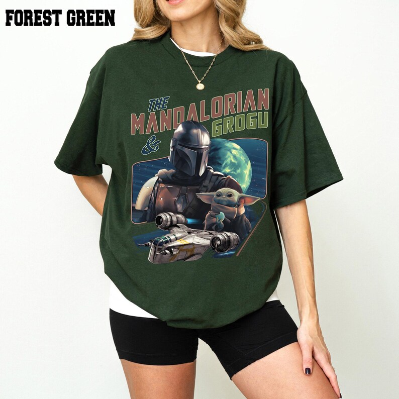 May include: Forest green t-shirt featuring a graphic of The Mandalorian and Grogu, with a spaceship and a planet in the background. The text "THE MANDALORIAN & GROGU" is printed above the graphic. The shirt is a dark green color.
