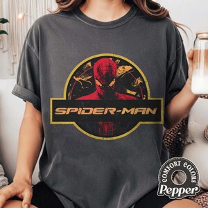 May include: A charcoal grey short-sleeve t-shirt featuring a graphic print of Spider-Man inside a yellow circular border. The design includes the text SPIDER-MAN in a bold yellow font across a horizontal black bar centred on the chest.