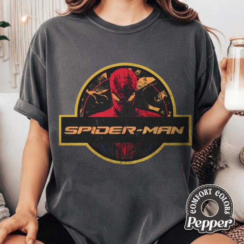 May include: A charcoal grey short-sleeve t-shirt featuring a graphic print of Spider-Man inside a yellow circular border. The design includes the text SPIDER-MAN in a bold yellow font across a horizontal black bar centred on the chest.