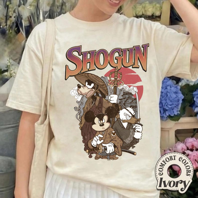 Puede incluir: Una camiseta de color marfil con un estampado gr&aacute;fico que incluye el texto SHOGUN en una fuente degradada. La ilustraci&oacute;n muestra a tres personajes de dibujos animados vestidos con atuendos japoneses tradicionales, incluyendo un sombrero c&oacute;nico.