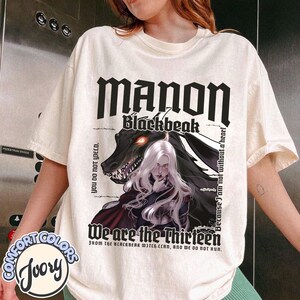 May include: Ivory Comfort Colors t-shirt featuring the text "MANON Blackbeak" in a gothic font, with an illustration of a woman with white hair and a dragon. The shirt also includes the phrases "We are the Thirteen" and "Because I am not without a heart."