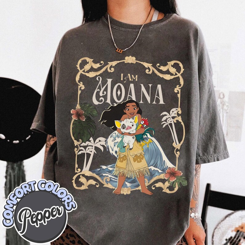 May include: A charcoal gray t-shirt featuring a graphic print of the character Moana holding a pig and a rooster. The design includes the text I AM MOANA, surrounded by a decorative gold frame with palm trees, hibiscus flowers, and ocean waves.