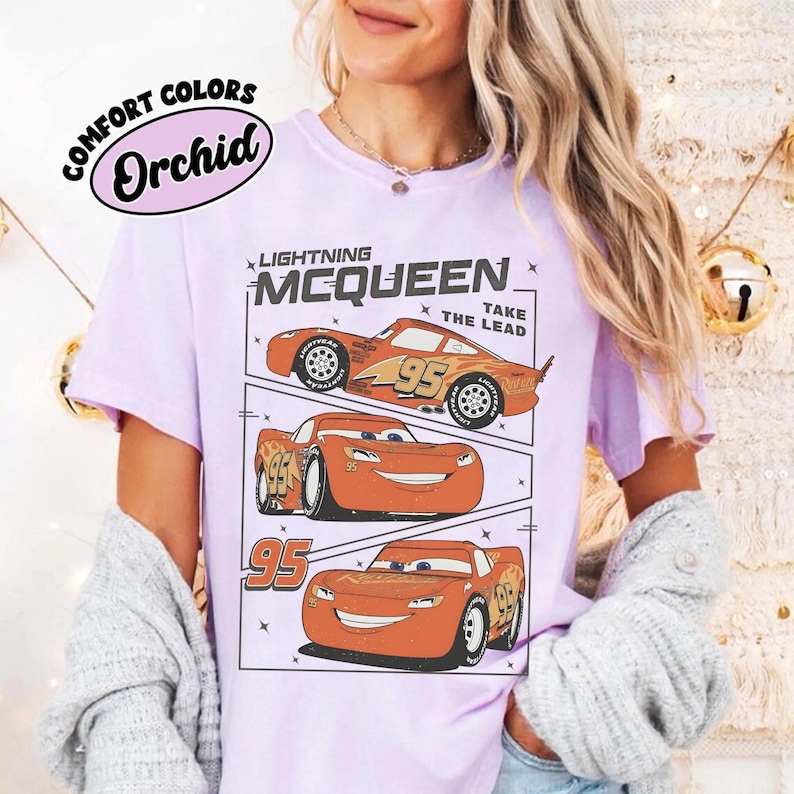 May include: A light purple t-shirt featuring a graphic of Lightning McQueen from the movie Cars. The design includes three images of the car with the number 95, and the text "LIGHTNING MCQUEEN TAKE THE LEAD". The shirt also has the text "COMFORT COLORS Orchid".
