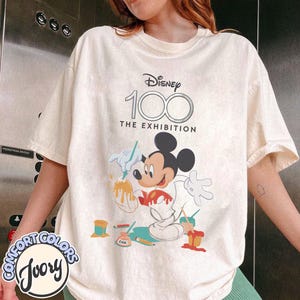 May include: Ivory-colored t-shirt with a graphic of Mickey Mouse painting, with the text "Disney 100 The Exhibition." The shirt has a crew neck and short sleeves. The Comfort Colors logo is visible on the bottom left.