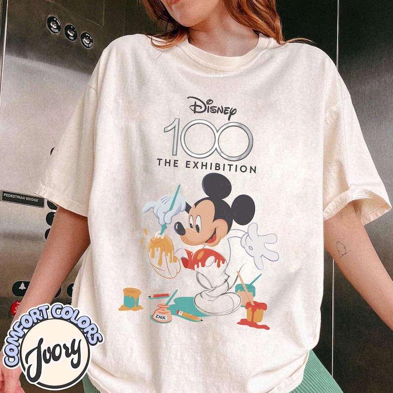 May include: Ivory-colored t-shirt with a graphic of Mickey Mouse painting, with the text "Disney 100 The Exhibition." The shirt has a crew neck and short sleeves. The Comfort Colors logo is visible on the bottom left.