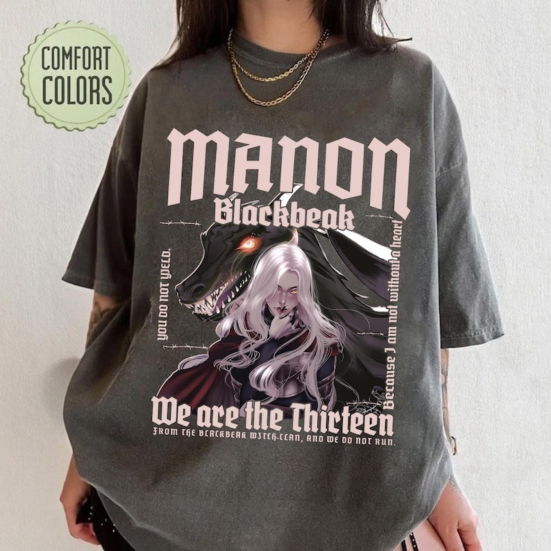 May include: Dark gray t-shirt featuring the text "MANON Blackbeak" above an illustration of a woman with white hair and a dragon. The shirt also includes the phrases "We are the Thirteen" and "You do not yield."