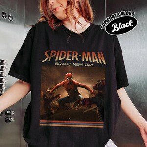 May include: A black short-sleeve t-shirt featuring a graphic print of Spider-Man in a dynamic pose. The text reads "SPIDER-MAN" and "BRAND NEW DAY" above the character. A small oval badge in the top right corner displays the text "COMFORT COLORS Black".