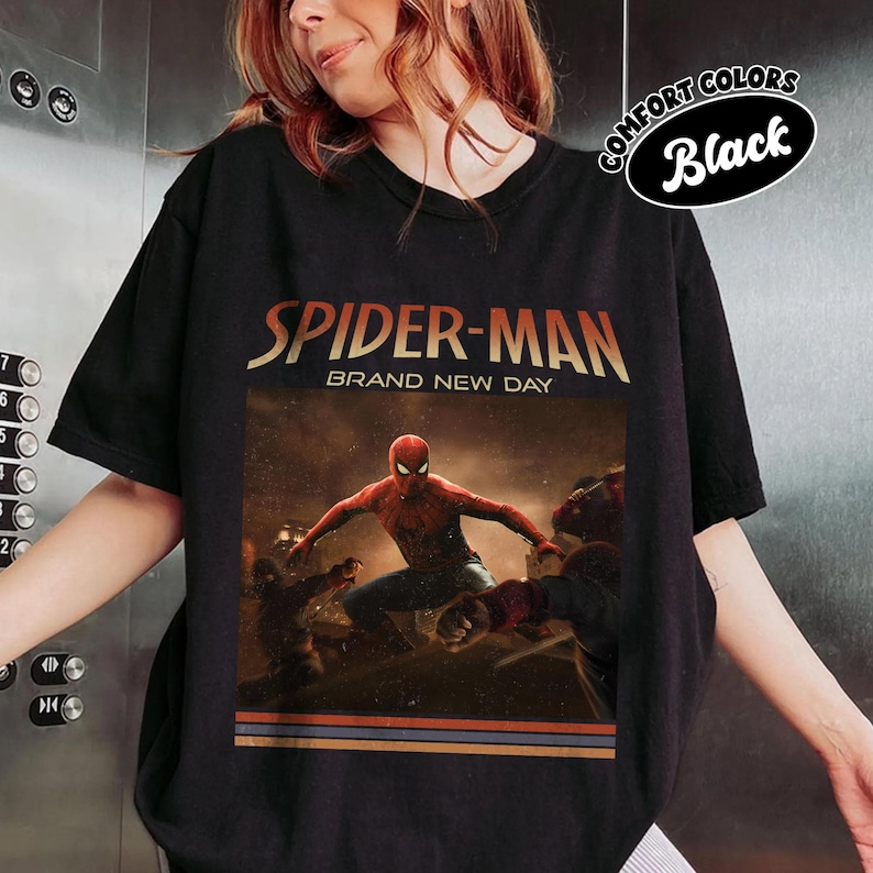 May include: A black short-sleeve t-shirt featuring a graphic print of Spider-Man in a dynamic pose. The text reads "SPIDER-MAN" and "BRAND NEW DAY" above the character. A small oval badge in the top right corner displays the text "COMFORT COLORS Black".