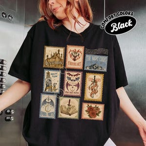 May include: A black t-shirt featuring a grid of nine rectangular postage stamp-style illustrations. The designs include a castle, a deer head, a mountain, an easel, bat wings, a sword, a crescent moon, an open book with a dagger, and red roses.
